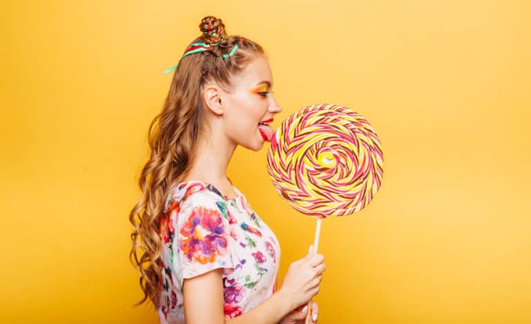 Beautiful young woman addicted to sugar with playful look eating huge candy and smiling. Stylish girl with blonde curly hair. Portrait of attractive lady with big lollypop, yellow wall on background.