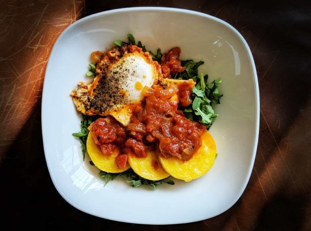 One white plate viewed from above containing one cooked egg, three slices of polenta, red marinara sauce, and some pops of green arugula.